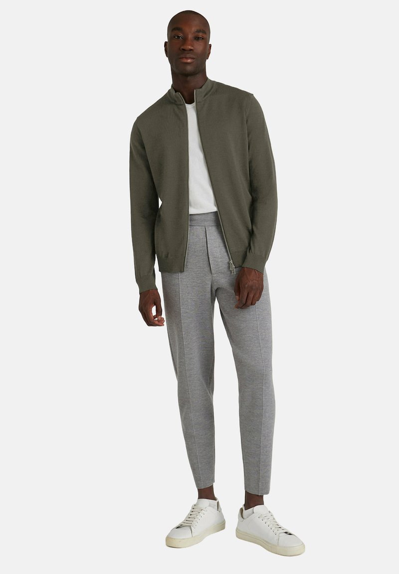 Olivengrøn zip-up jakke over en hvid T-shirt, parret med grå skræddersyede sweatpants og hvide sneakers. Glat tekstur, afslappet design.