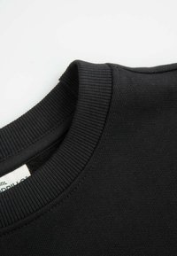 Czarny sweter z okrągłym dekoltem, z teksturą ściągacza i częściowo widoczną białą metką odzieżową wewnątrz.