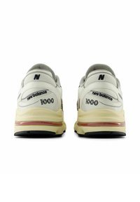 Weiße und beige New Balance Sneakers mit einer gepolsterten Sohle, ausgestattet mit schwarzem Branding, strukturiertem Obermaterial und reflektierenden Akzenten.