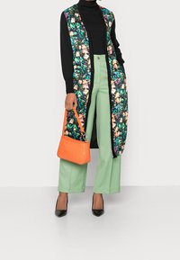 Bloemenlange cardigan met een zwarte achtergrond, zwarte coltrui, mintgroene wijde broek en een oranje handtas. Zwarte spitse hakken.