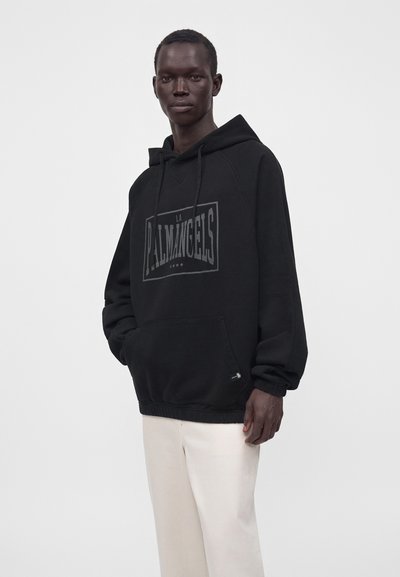 Άνδρας που φοράει μαύρο φαρδύ hoodie με κείμενο "LA PALMANGELS" και παντελόνι κρεμ, στέκεται μπροστά σε απλό ανοιχτόχρωμο φόντο.