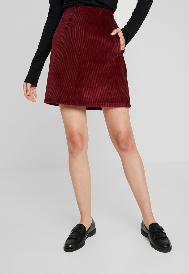 Donna che indossa una gonna rossa in velluto a coste con tasche, un top a maniche lunghe nero e mocassini neri con frange, in piedi davanti a uno sfondo grigio uniforme.
