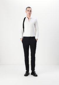 Weißes Hemd mit Button-Down-Kragen und Slim-Fit, schwarze maßgeschneiderte Hose und schwarze Schuhe. Ein schwarzer Träger liegt lässig über einer Schulter.