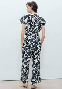 Combinaison florale en crème avec un motif de feuilles bleu marine, manches courtes à volants, taille nouée et dos ouvert. Tissu doux et léger.