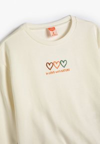 Sudadera de color crema con un cuello acanalado. Presenta un mensaje bordado "en AMOR con la NATURALEZA" y tres diseños de corazones en marrón, naranja y verde.