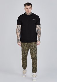 Čierne tričko s krátkym rukávom s malým logom, v kombinácii so zelenými maskáčovými teplákmi s vreckami a elastickými manžetami, doplnené o biele tenisky.