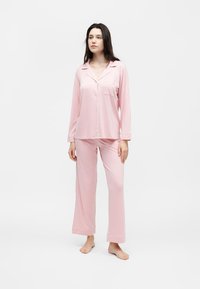 Γυναίκα στεκόμενη ξυπόλυτη, ντυμένη με ανοιχτό ροζ μακρυμάνικο πουκάμισο πιτζάμα με κουμπιά και χαλαρό παντελόνι, μπροστά σε απλό λευκό φόντο.