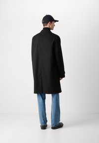 Manteau noir avec un col montant, des manches longues et des poignets boutonnés, associé à un jean bleu clair et des chaussures noires. Détails de couture visibles.