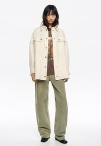 Camisa chaqueta de lana mezclada en color crema, de corte oversized, con botones marrones y dos bolsillos en el pecho, combinada con pantalones de pana anchos en verde oliva y botas negras de tobillo.
