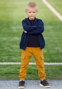 Dziecko w granatowej kurtce na suwak, czarnej koszulce, żółtych spodniach cargo i czarnych butach, stojące z założonymi rękami na tle zielonego pola.