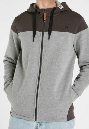 Mann trägt eine zweifarbige, grau-dunkelbraune Kapuzenjacke mit Reißverschluss, schwarzen Kordeln und Seitentaschen, kombiniert mit hellblauen Jeans.