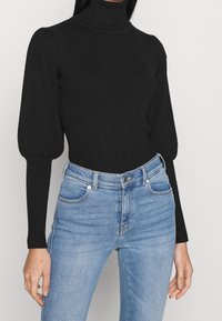 Zwarte ribbel turtleneck trui met opgezette mouwen, gedragen met lichtblauwe skinny jeans; gladde textuur en subtiele stretch voor comfort.