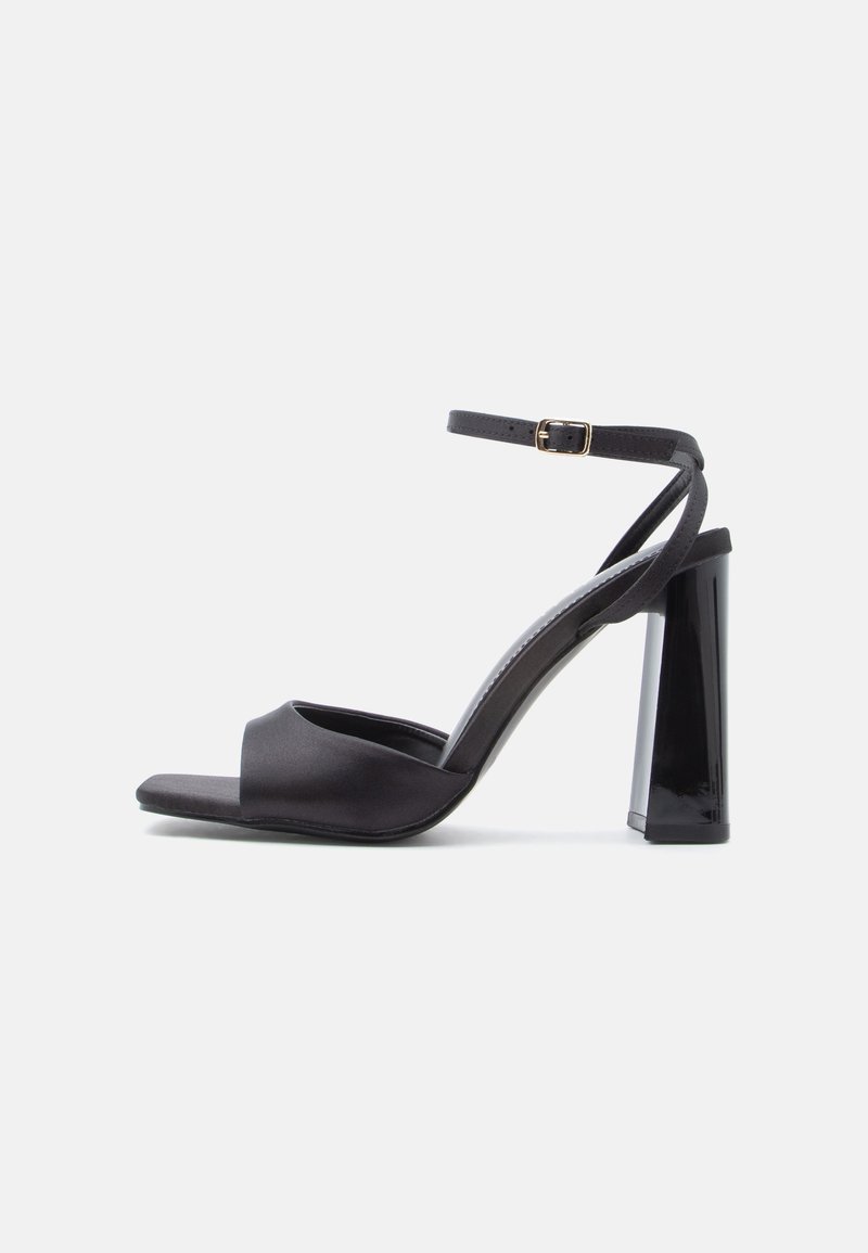 Melni satīna augstpapēžu sandales ar potītes siksniņu, atvērtu purngalu un spīdīgu bloka papēdi. Raksturo minimālas šuves un gludu tekstūru.
