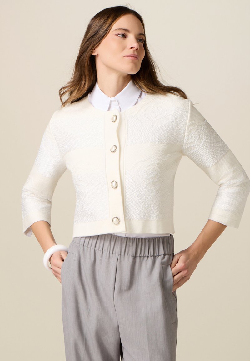 Donna che indossa un cardigan corto bianco con trama e bottoni di perle sopra una camicia bianca con colletto e pantaloni grigi larghi, con le mani nelle tasche.