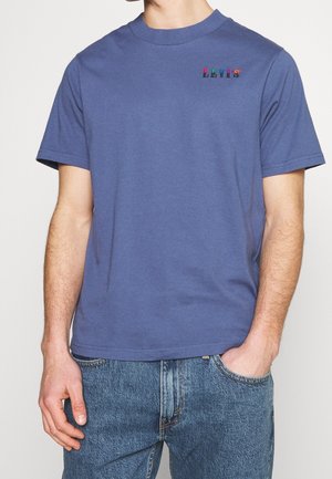 Mężczyzna w niebieskim krótkim rękawem T-shircie z kolorowym logo „LEVI'S” na klatce piersiowej i niebieskich dżinsach, z jedną ręką w kieszeni.