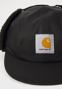 Cappello nero realizzato in nylon testurizzato con visiera piatta, dotato di un'etichetta quadrata bianca con un logo giallo e il testo "carhartt."