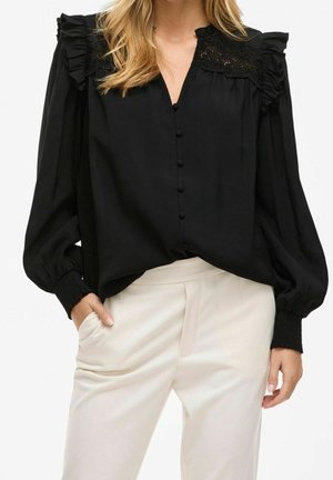 Nainen pukeutuneena mustaan pitkähihaiseen paitapuseroon, jossa on ruchesomisteiset hartiat, ja valkoisiin korkeavyötäröisiin housuihin, poseeraa käsi taskussa.