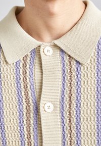 Gebreid poloshirt in beige met getextureerde verticale strepen in lavendel, lichtbruin en crème. Voorzien van een klassieke kraag en twee knopen.