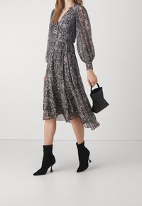 Robe fluide avec un motif serpent noir et gris, manches longues transparentes, taille cintrée, jupe superposée, et associée à des bottines noires et un mini sac.