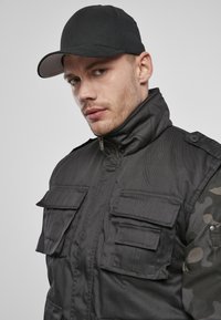 Svart polstret jakke med flere frontlommer, høy krage og camo-ermer; kombinert med en svart caps med buet skjerm.