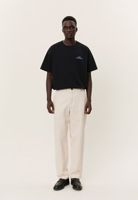 Mlad moški, ki nosi črno majico s napisom "Vstop prost", bež hlače in črne čevlje, stoji pred enobarvnim svetlim ozadjem.