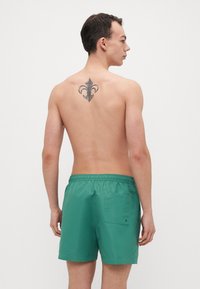 Grønne badeshorts laget av lett stoff, med elastisk midje og en enkelt baklomme. Enkel design med glatt tekstur.