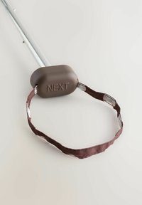 Hnědá gumová rukojeť s nápisem "NEXT" a texturovaným úchopem, připevněná k stříbrnému kovovému sloupu a tmavě hnědému popruhu s jasnými akcenty.