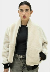 Cremfarbener Kunstpelz-Bomberjacke mit schwarzer Lederfront, gerippten Bündchen und Reißverschluss. Getragen über einem schwarzen gerippten Oberteil und Jeans.