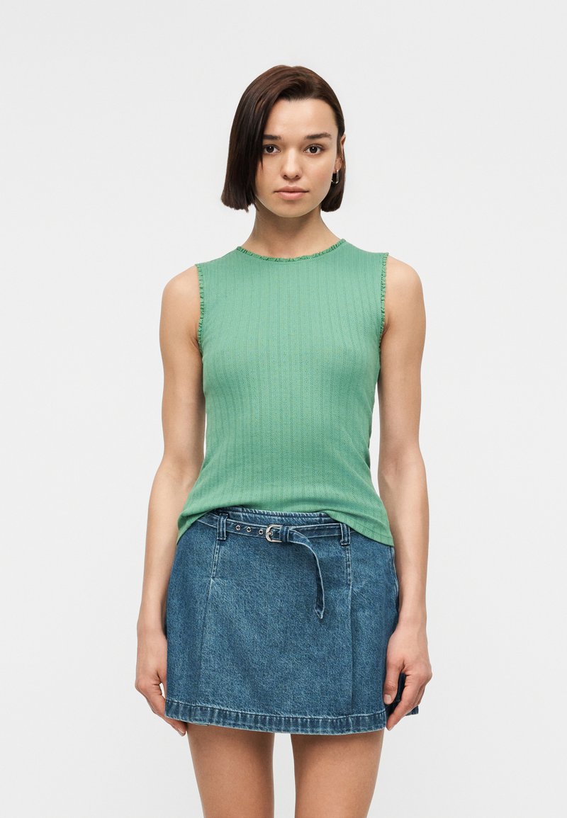 Femme aux cheveux bruns courts portant un haut côtelé vert sans manches et une mini-jupe en denim bleue ceinturée, debout devant un fond blanc.