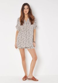 Kurzarm-Tunika mit gemustertem, geometrischem Design in Beige und Braun. Weicher Stoff, lockere Passform, endet oberhalb des Oberschenkels. Getragen mit braunen Rutsch-Sandalen.