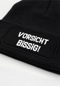 Black knitted beanie with a textured finish featuring a white rectangular patch that reads "VORSICHT BISSIG!" in bold letters.