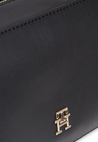 Musta nahkalaukku, jossa on teksturoitu pinta ja kullanvärinen logoetuliite edessä. Kompakti muotoilu, jossa on kestävä ompelu.