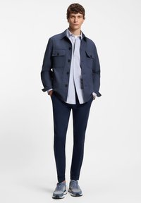 Tamsiai mėlynas švarkas su keturiomis priekinėmis kišenėmis, derinamas su šviesiai mėlyna marškinėliais ir tamsiai mėlynomis siaurėjančiomis kelnėmis. Sportbačiai pasižymi mėlyna ir pilka spalvų schema.