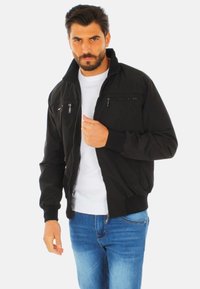 Giubbotto bomber nero con zip frontale, polsini e orlo a costine. Presenta due tasche sul petto con zip e una finitura liscia. Indossato sopra una camicia bianca.