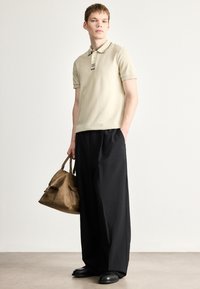 Μπεζ polo με κοντά μανίκια και σκούρες λεπτομέρειες, μαύροι φαρδιοί παντελόνες και καφέ δερμάτινη τσάντα, σε φόντο ανοιχτό γκρι.