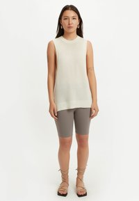 Kvinna iklädd ärmlös vit stickad topp, grå åtsittande knälånga shorts och beige sandaletter med snörning, stående mot vit bakgrund.