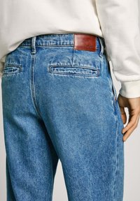 Ljusblå denimjeans med en något blekt textur, med en brun läderlappar bak och två bakfickor, kombinerat med en vit tröja.