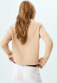 Svetle béžový pletený svetr s dlouhými rukávy, žebrovanými manžetami a lemem; hladká textura a zkrácený design. Zádní pohled, spárovaný s bílými kalhotami.