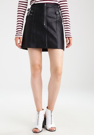 Sort lædernederdel i minilængde med lynlås foran og bukseseler i siden, båret med en langærmet top med striber og hvide åbne hæle med peep-toe.