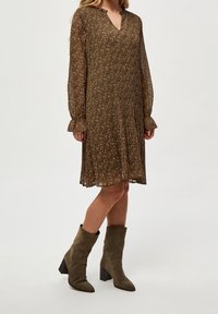 Ruskea kukkamekko, jossa on pitkät hihat, V-pääntie, röyhelömanchetit ja levenevä hame. Yhdistetty polvitason oliivinvihreisiin mokkasaappaisiin.