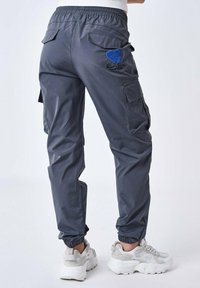 Pantaloni cargo grigi con cintura elasticizzata, tasche laterali e posteriormente, caratterizzati da un design a cuore blu e dettagli ricamati. Abbinati a sneakers bianche.