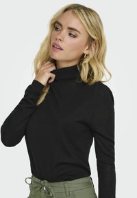 Schwarzer Langarm-Turtleneck-Pullover mit körpernahem Design, weicher Textur und Seitennähten, kombiniert mit olivgrünen, hochtaillierten Hosen.