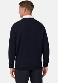 Tamsiai mėlynas megztinis su ribuotais rankogaliais ir apatine dalimi, pasižymintis lygiu tekstūru ir apvaliu iškirpimu. Šiek tiek laisvas pritaikymas, vientisas dizainas.