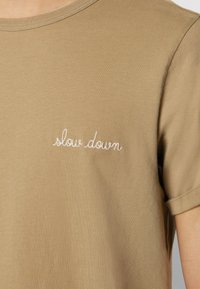 T-shirt à manches courtes en lin avec une broderie cursive blanche "ralentis" sur le côté gauche de la poitrine.