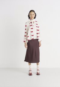 Blouse crème avec des motifs de lèvres rouges, associée à une jupe midi marron. Les accessoires incluent des chaussettes blanches avec des lèvres rouges et des chaussures brillantes marron.