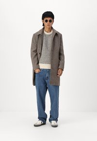 Cappotto di lana marrone, maglione a maglia con motivo, jeans in denim blu larghi e sneakers bianche. Gli occhiali da sole completano il look.