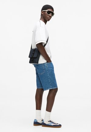 Tricou alb supradimensionat, pantaloni scurți din denim albastru, geantă neagră purtată în diagonală, ochelari de soare albi și adidași albaștri cu accente crem și talpă maro.