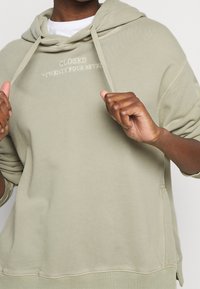Sudadera con capucha verde de corte relajado, con un bolsillo canguro y texto bordado "CERRADO 'VEINTICUATRO SIETE'" en el pecho.