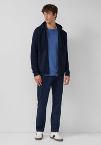 Jonge man in een marineblauwe rits-hoodie, blauwe t-shirt, donkerblauwe broek en witte sneakers, staande tegen een effen grijze achtergrond.