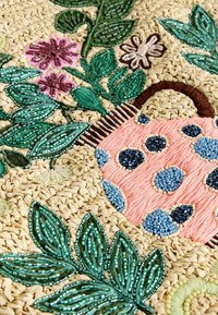 Pärlat konstverk med en rosa korg med blå prickar, omgiven av gröna blad och blommor i rosa, vitt och brunt på en halm bakgrund.